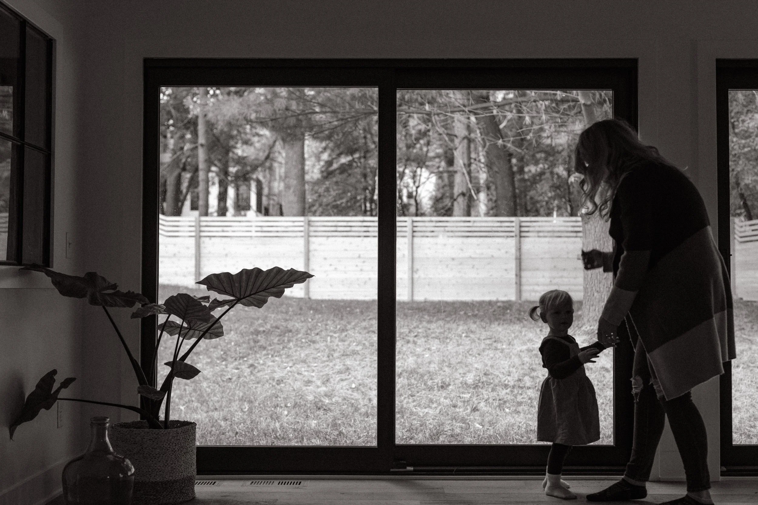 A black-and-white photo of a woman and a young girl near a large sliding glass door leading to a backyard with trees and a wooden fence. Inside, potted plants are visible.