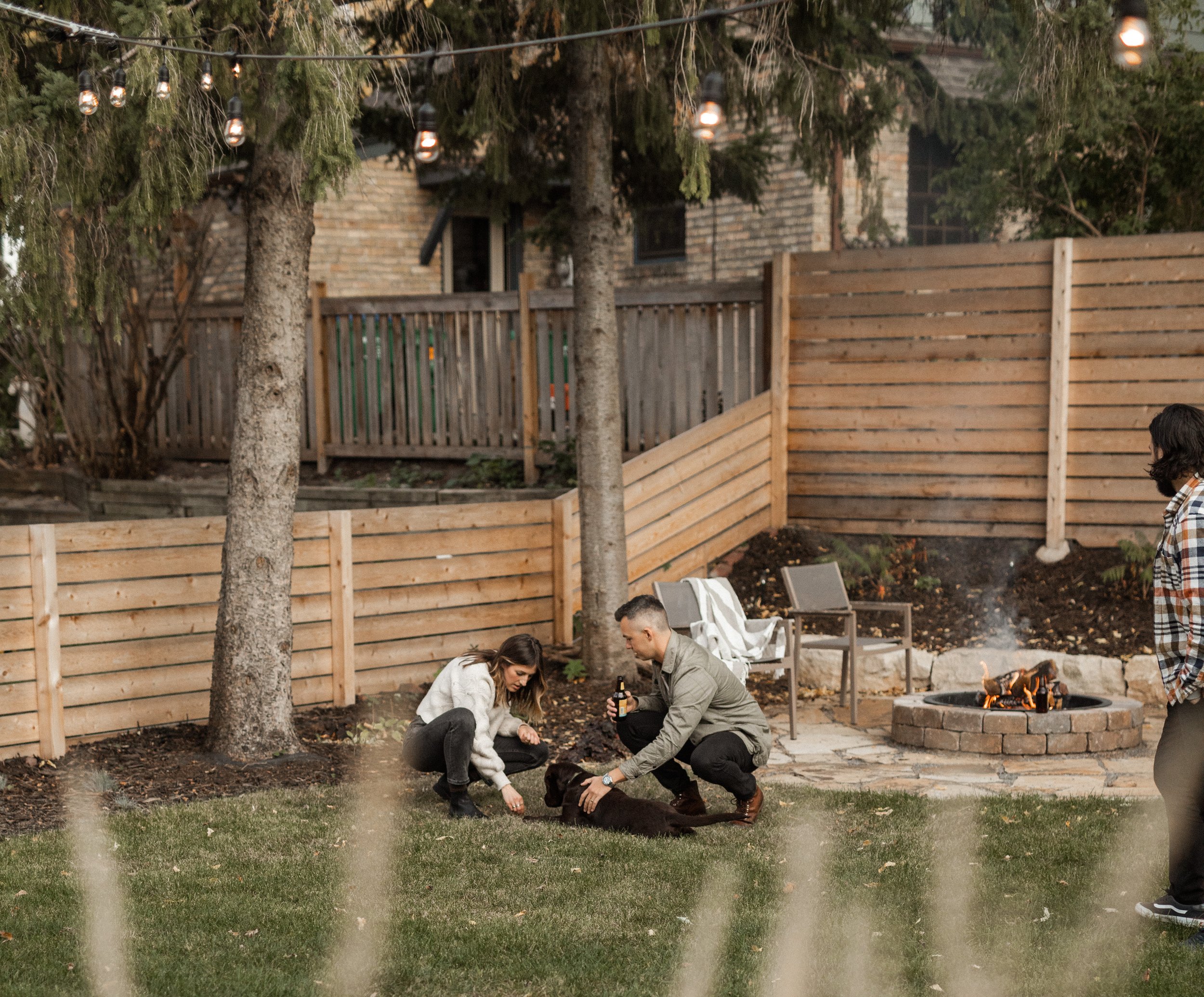 People gathered around a small fire pit in a backyard, with a wooden fence and trees in the background, under string lights hanging from the trees.