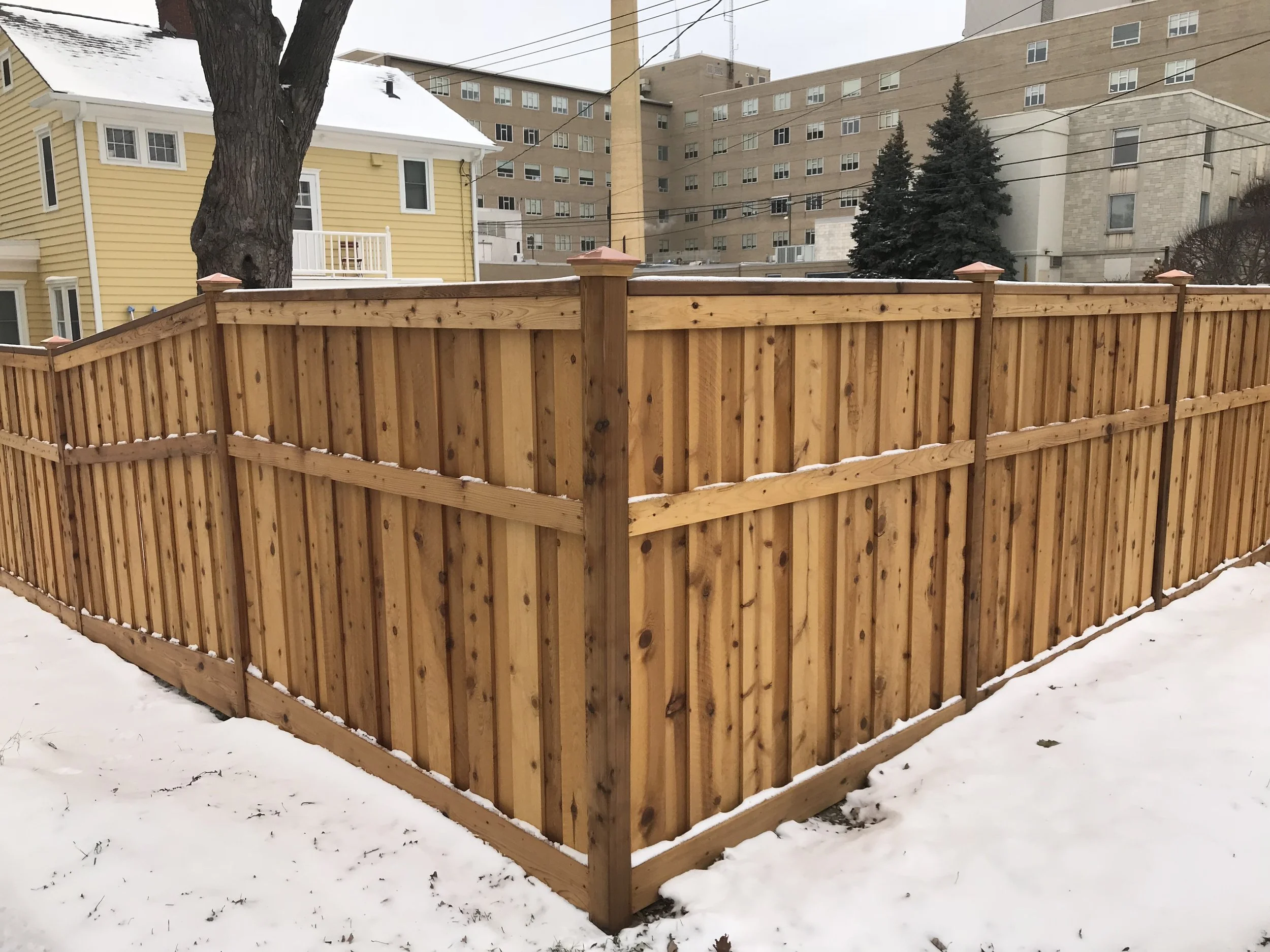 A wooden privacy fence surrounds a snow-covered yard, with a large tree and neighboring buildings in the background.