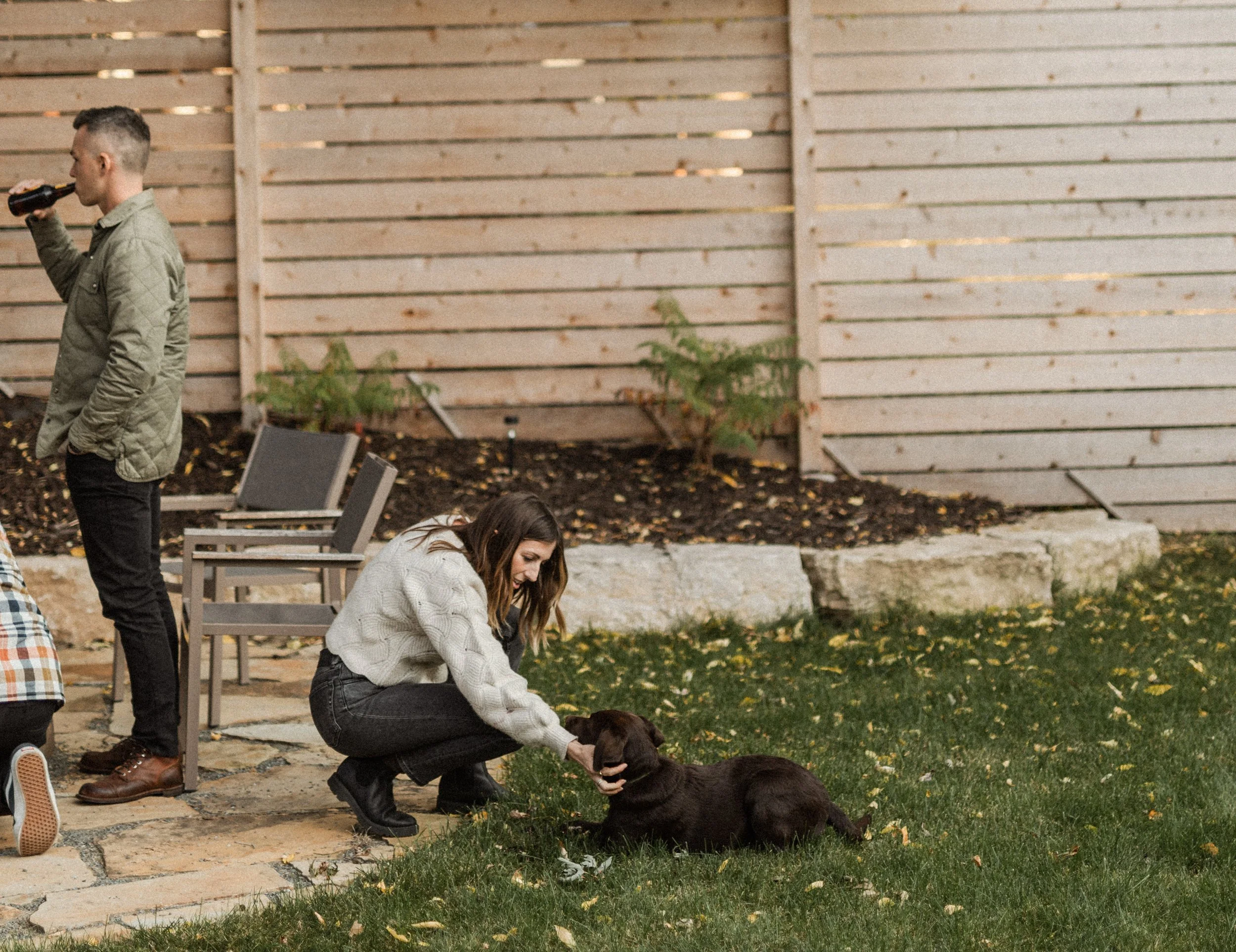 A young woman in a gray sweater and black jeans is kneeling on grass, petting a brown puppy. Another woman in a green jacket and black pants is standing nearby, drinking from a bottle. There are wooden benches and a wooden fence in the background, wi