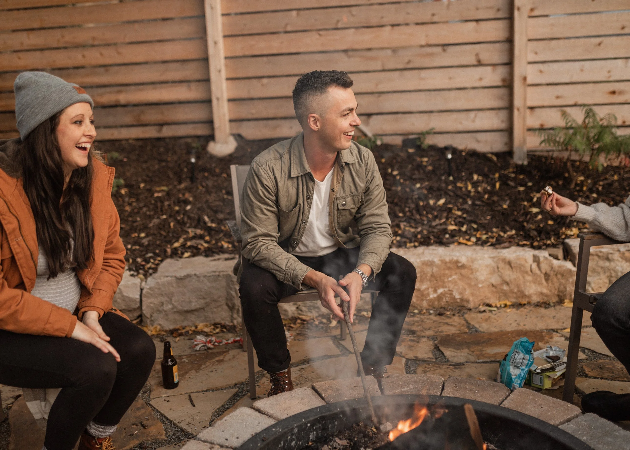 Three people sitting outdoors near a fire pit, smiling and socializing, with a wooden fence and some plants in the background.