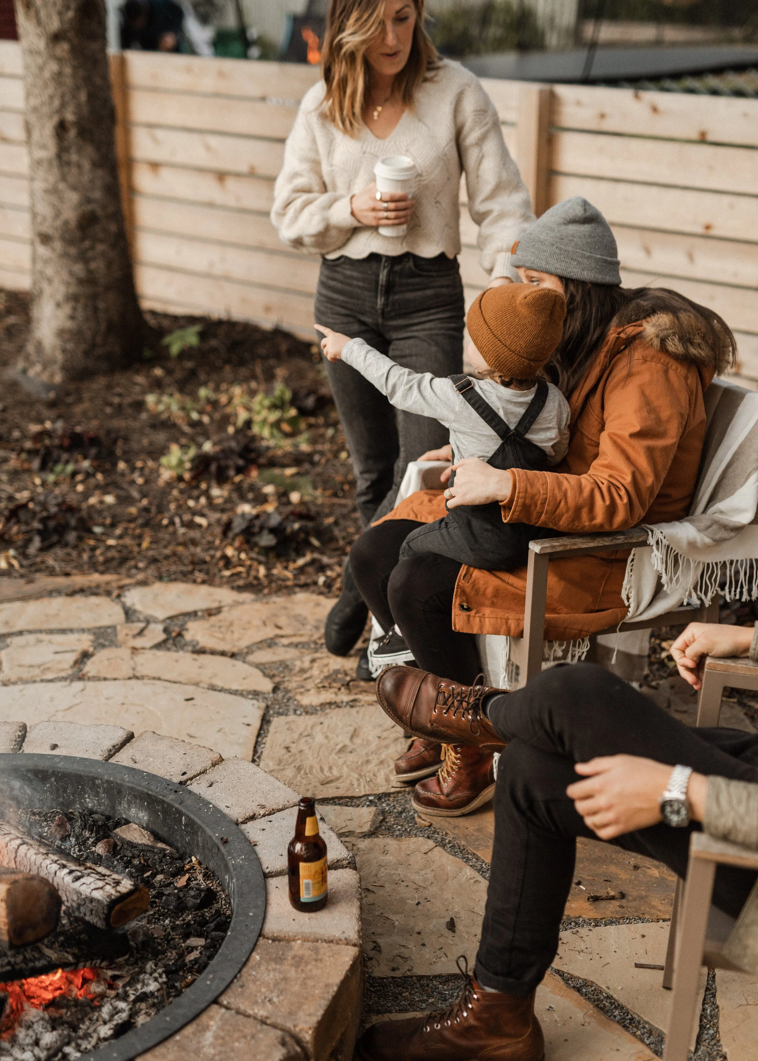 A woman is standing near a group of people, holding a cup of coffee, while a woman with long dark hair and a gray beanie, wearing a brown jacket, and a child wearing a brown beanie and a gray shirt with suspenders, are sitting on a bench and talking 