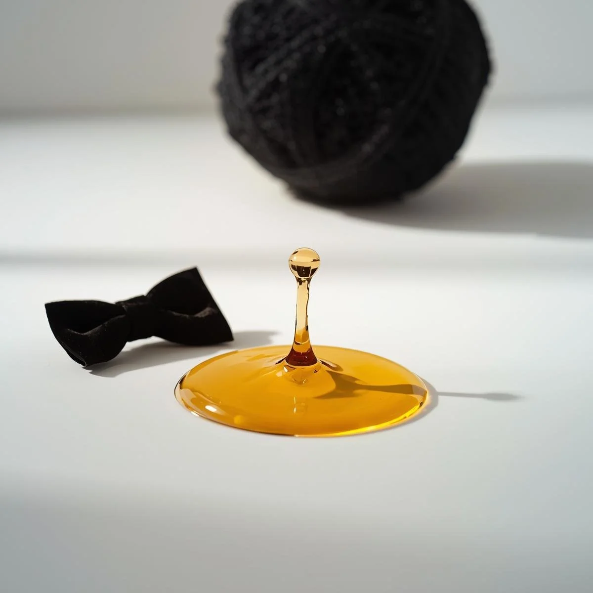 A viscous, translucent amber body wash droplet cascades in slow motion next to a single, a black velvet bow tie on a clean, minimalist white surface. A textured, black mesh shower sponge is softly blurred in .jpg