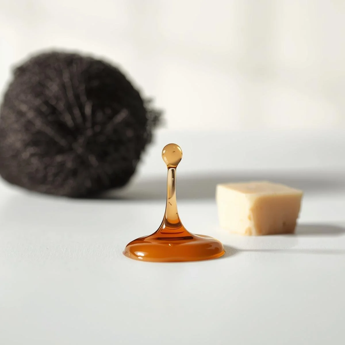A minimalist and clean photography style. A viscous, translucent amber body wash droplet cascades in slow motion next to a single. Cocoa Butter Chunk on a clean, white surface. A textured, black mesh shower s.jpg