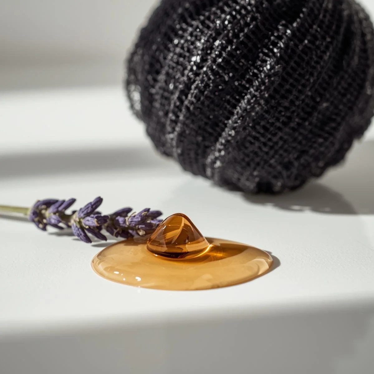 A minimalist and clean photography style. A viscous, translucent amber body wash droplet cascades in slow motion next to a single. Lavender sprig on a clean, white surface. A textured, black mesh shower spong.jpg