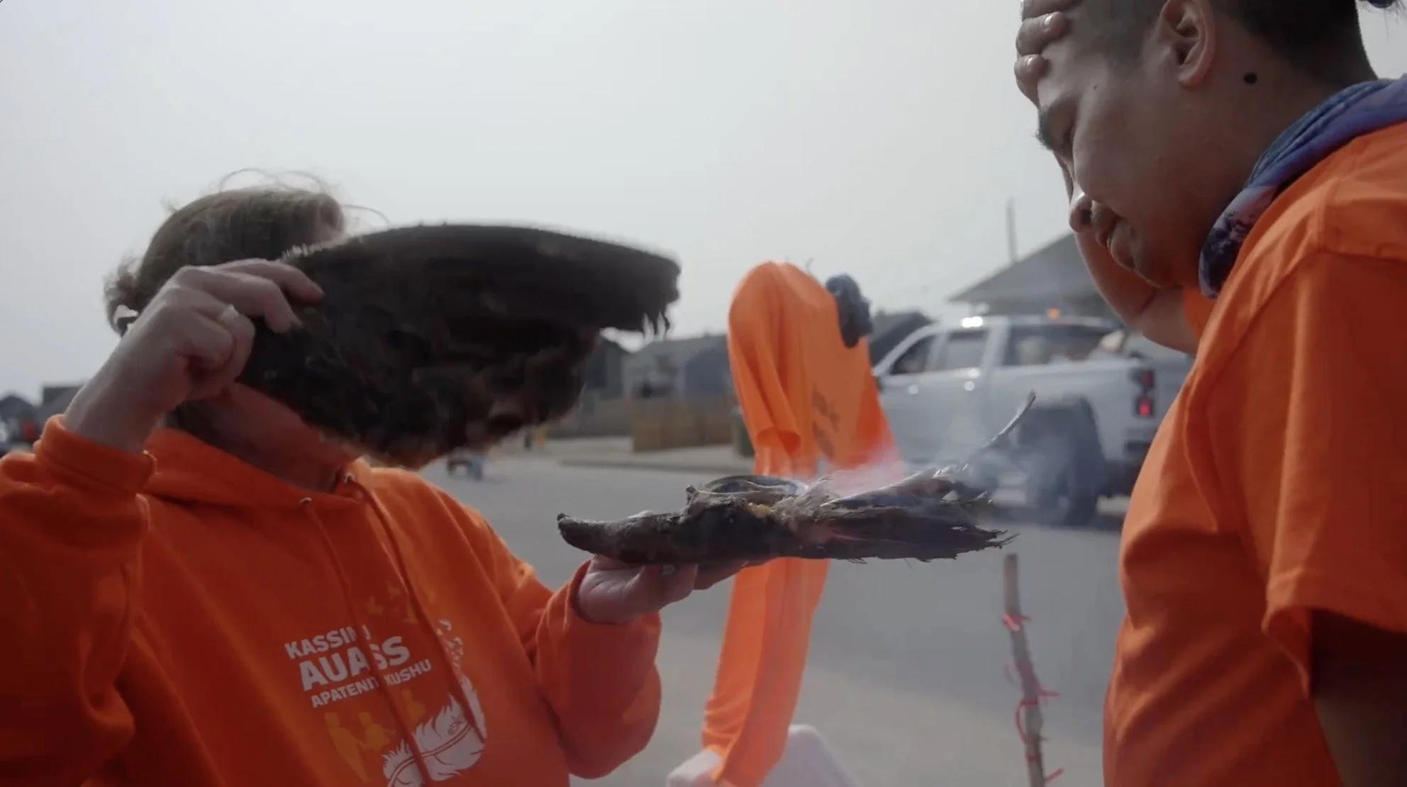 Deux personnes en vestes orange discutent près d'une voiture, une d'elles tient un poisson fumé, dans un environnement extérieur.