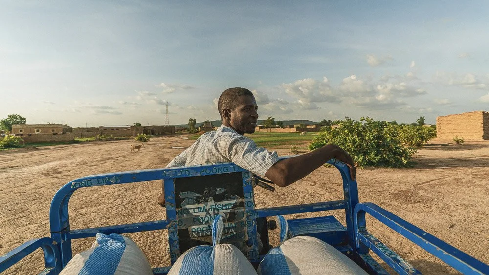 Un homme assis à l'arrière d'un vélo ou d'un chariot dans un paysage rural, avec des bâtiments en terre cuite et des buissons au loin.