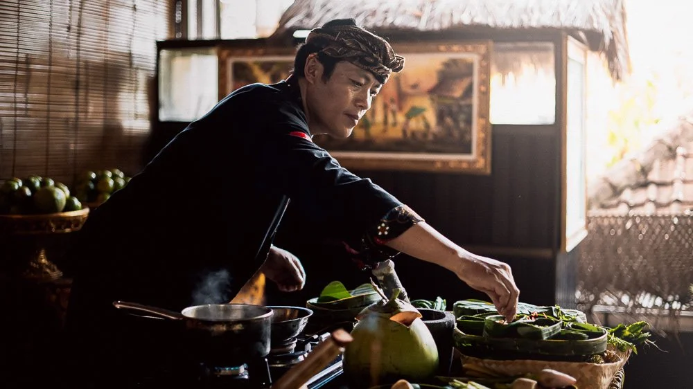 Un homme prépare un repas Balinais dans une cuisine en bois, entourée de paniers avec des fruits et des légumes, avec une lumière naturelle qui illumine la pièce.
