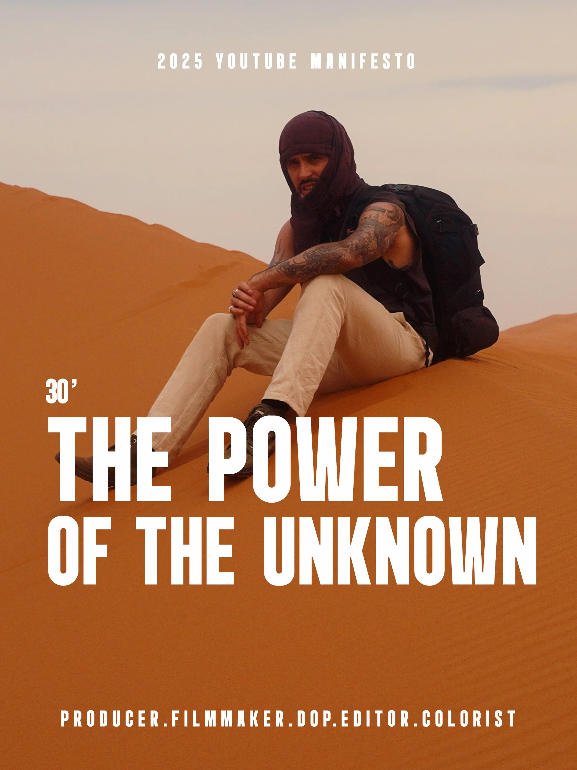 Un homme avec un sac à dos, des tatouages, portant un turban et des vêtements sombres, assis sur une dune de sable dans un désert au coucher du soleil.