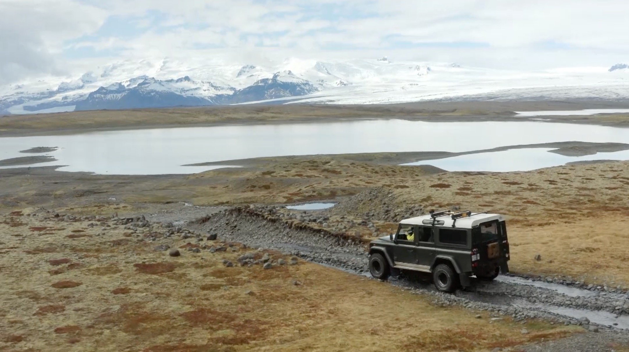 Un véhicule tout-terrain noir traverse un chemin rocailleux dans une région isolée avec des lacs et des montagnes enneigées au loin.