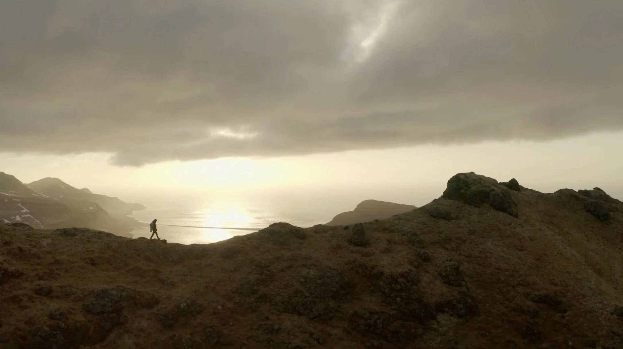 Un randonneur marche sur une crête rocheuse sous un ciel nuageux, avec une vue sur la mer et un horizon lumineux en arrière-plan.