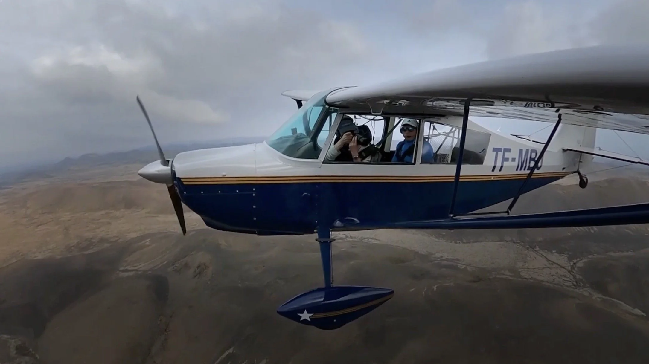 Avion de tourisme en vol au-dessus d'un paysage désertique avec deux personnes à bord, une avec un casque de pilote et l'autre avec des lunettes de soleil.