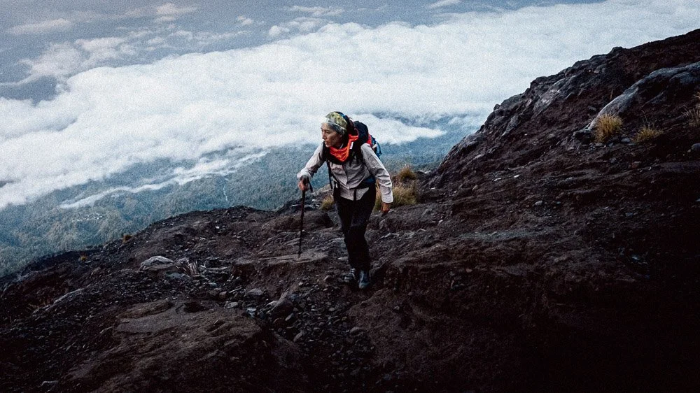 Femme faisant de la randonnée en montagne avec un bâton, portant un manteau et un sac à dos, chemin escarpé avec nuages en arrière-plan.
