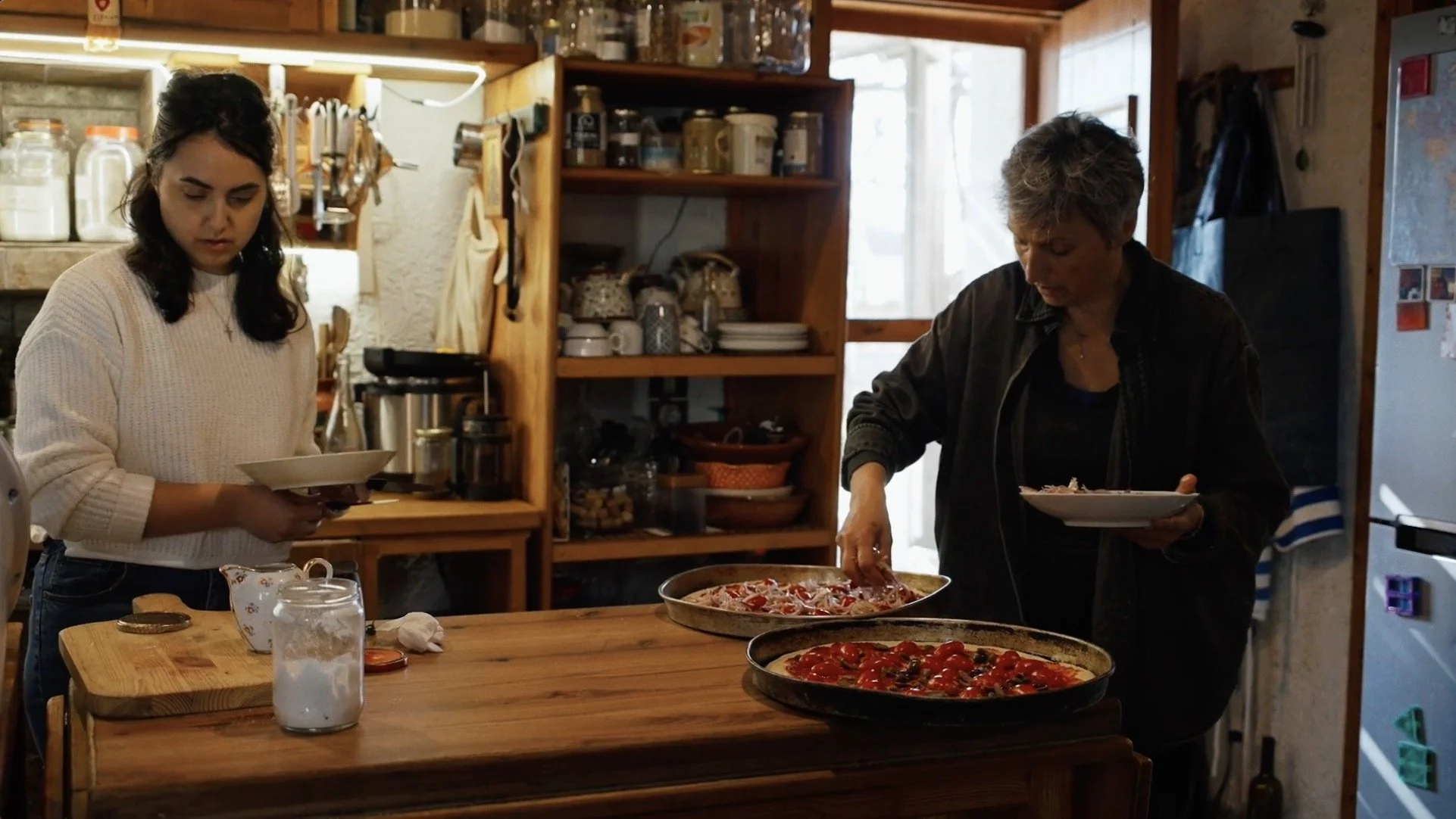 Une jeune femme et une femme plus âgée dans une cuisine rustique préparant ou servant des pizzas ou des tartes. La jeune femme tient une assiette blanche, la plus âgée manipule une pizza sur une plaque de cuisson.