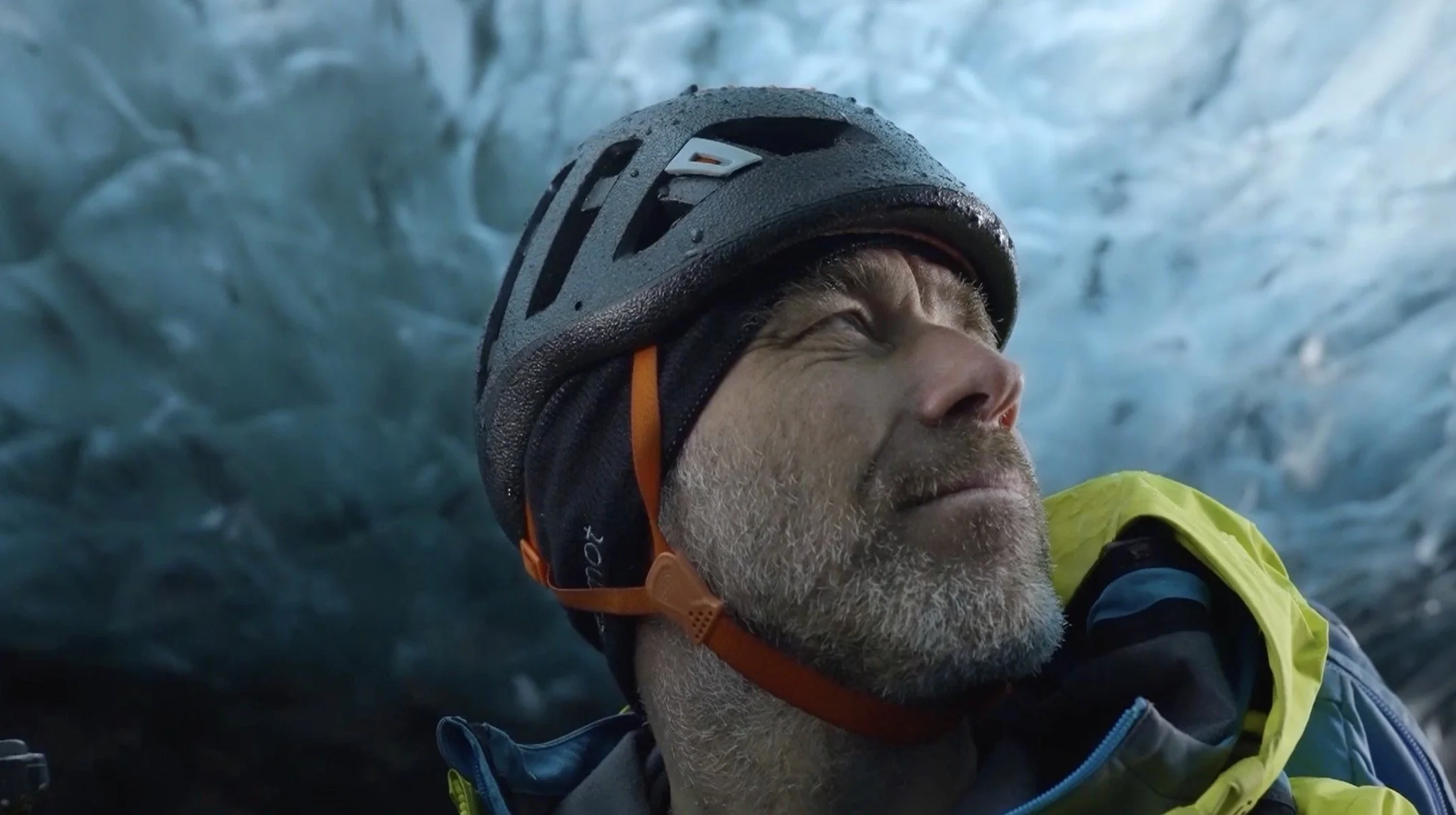 Un homme portant un casque d'escalade à l'intérieur d'une grotte de glace, souriant avec le regard vers le haut.