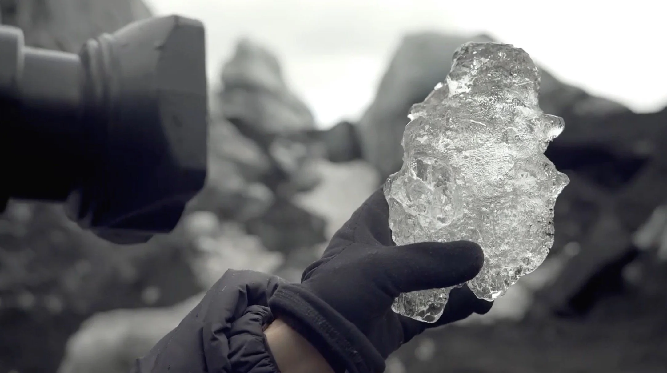 Personne tenant un morceau de glace dans une main gantée, avec un paysage rocheux en arrière-plan.