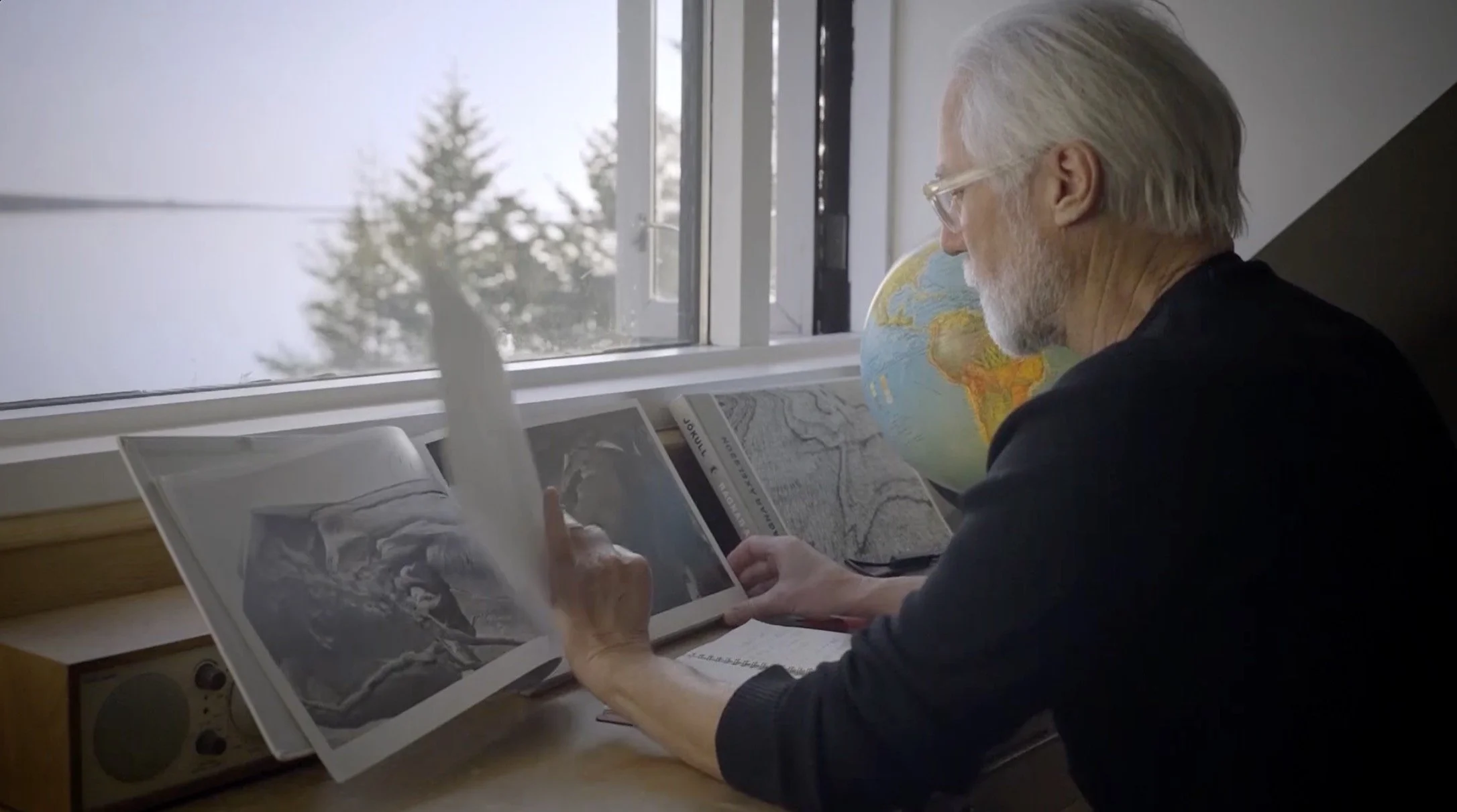 Un homme âgé avec des cheveux gris et une barbe, portant des lunettes, regarde des documents et des photos près d'une fenêtre, avec une globe terrestre et une carte à ses côtés.