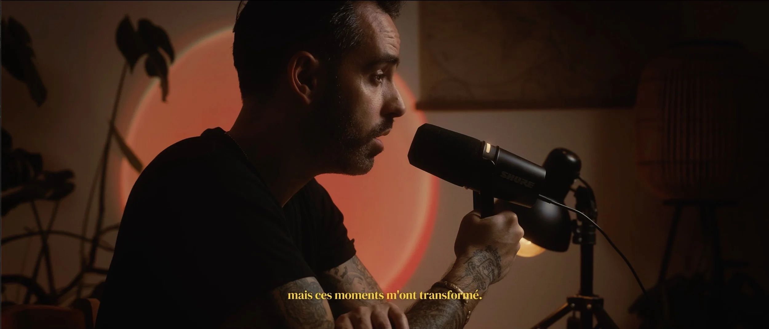 Un homme blond, tatoué, enregistrement dans une pièce sombre avec un microphone Shure, sous une lumière orange, avec des meubles en fond et une citation en français en sous-titre.