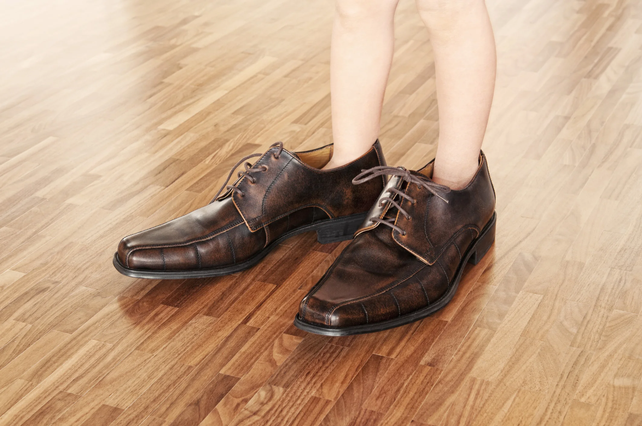 A child wearing a mans dress shoe on a wooden floor and only his legs being shown..