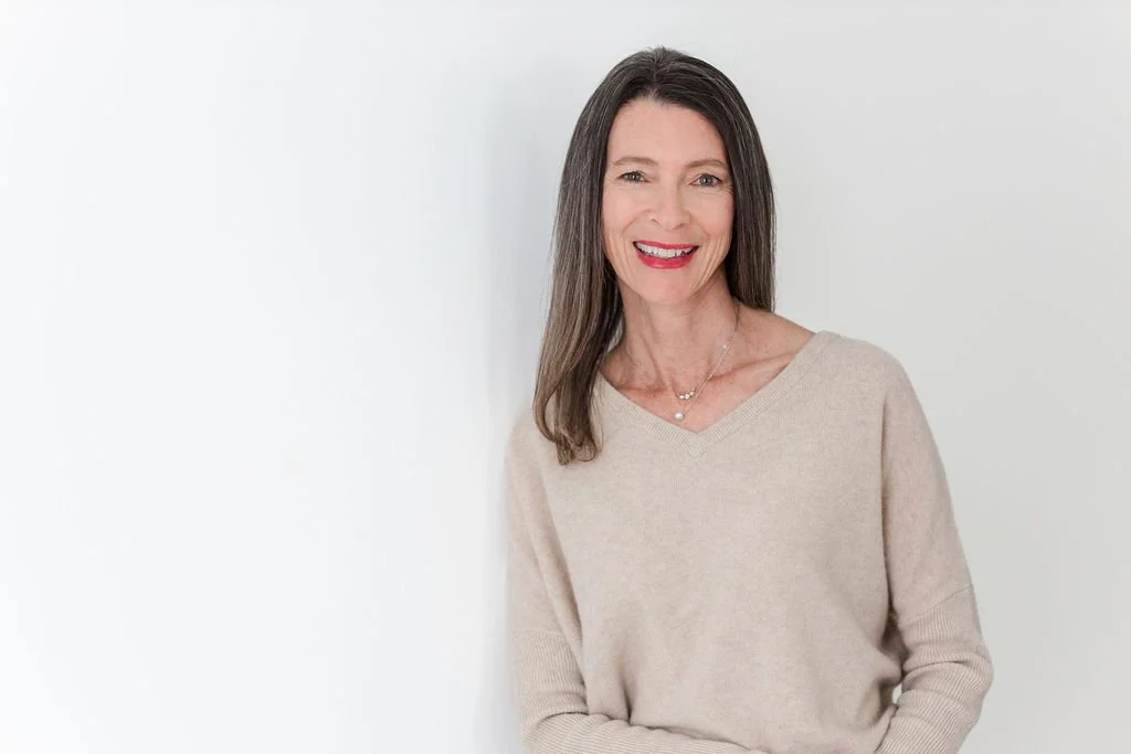 A woman with long dark hair wearing a beige sweater, smiling, standing against a white background.
