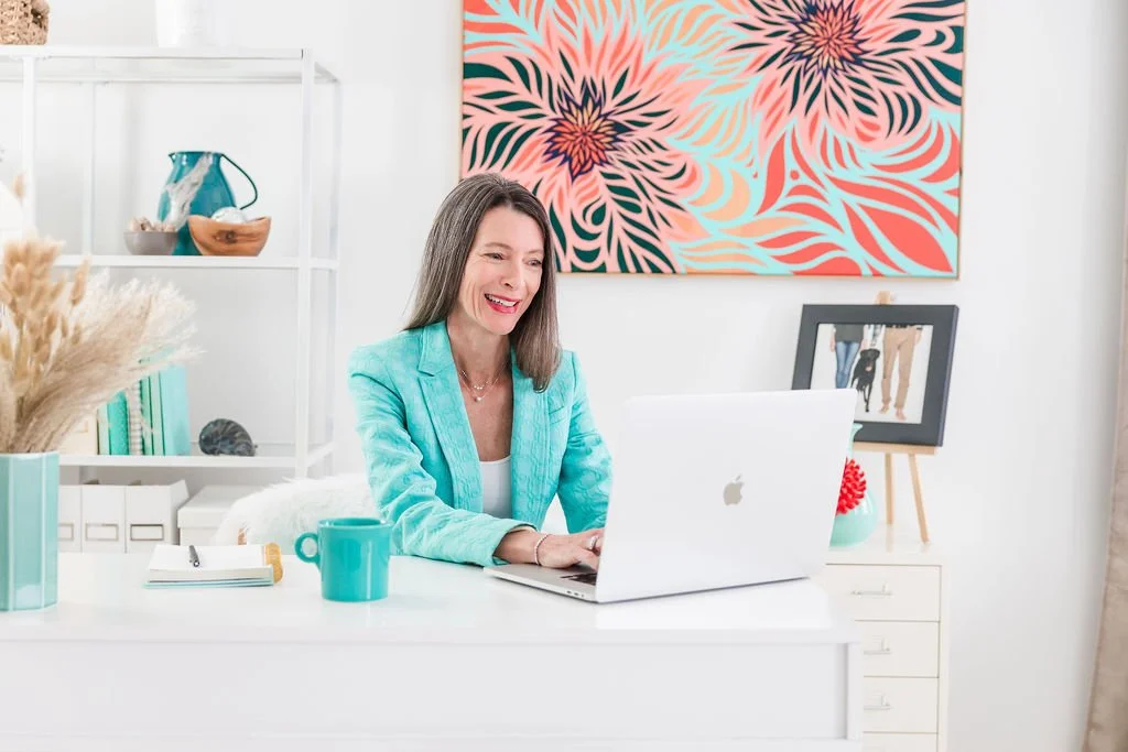 A woman working at a white desk with a silver laptop, a turquoise mug, and a notebook, in a bright and decorated home office.