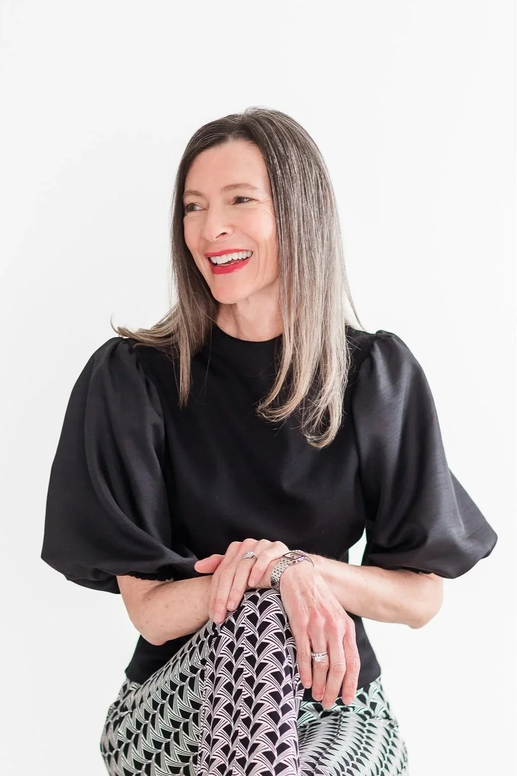 A woman with shoulder-length gray hair, wearing a black puff-sleeve top, smiling with red lipstick, sitting with her hands folded over her knee, in front of a plain white background.