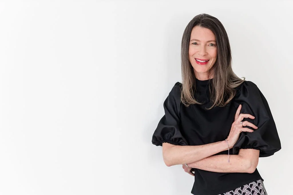 A woman with shoulder-length brown hair, smiling, wearing a black puff-sleeve top, and standing against a white background.