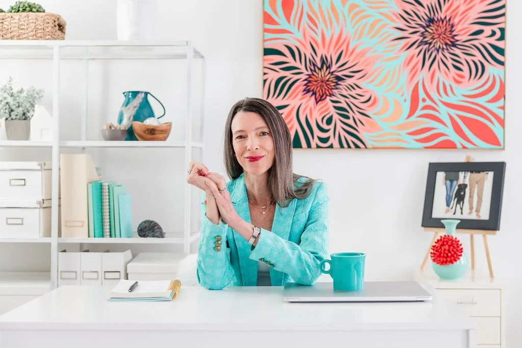 A woman with long dark hair smiling at a white desk in a modern office or home office. She is wearing a turquoise blazer and sitting with her hands clasped. On the desk are a teal mug, a closed laptop, and a notepad with a pen. Behind her are white shelves with decorative items, books, and storage boxes. A colorful, abstract floral art piece hangs on the wall behind her.