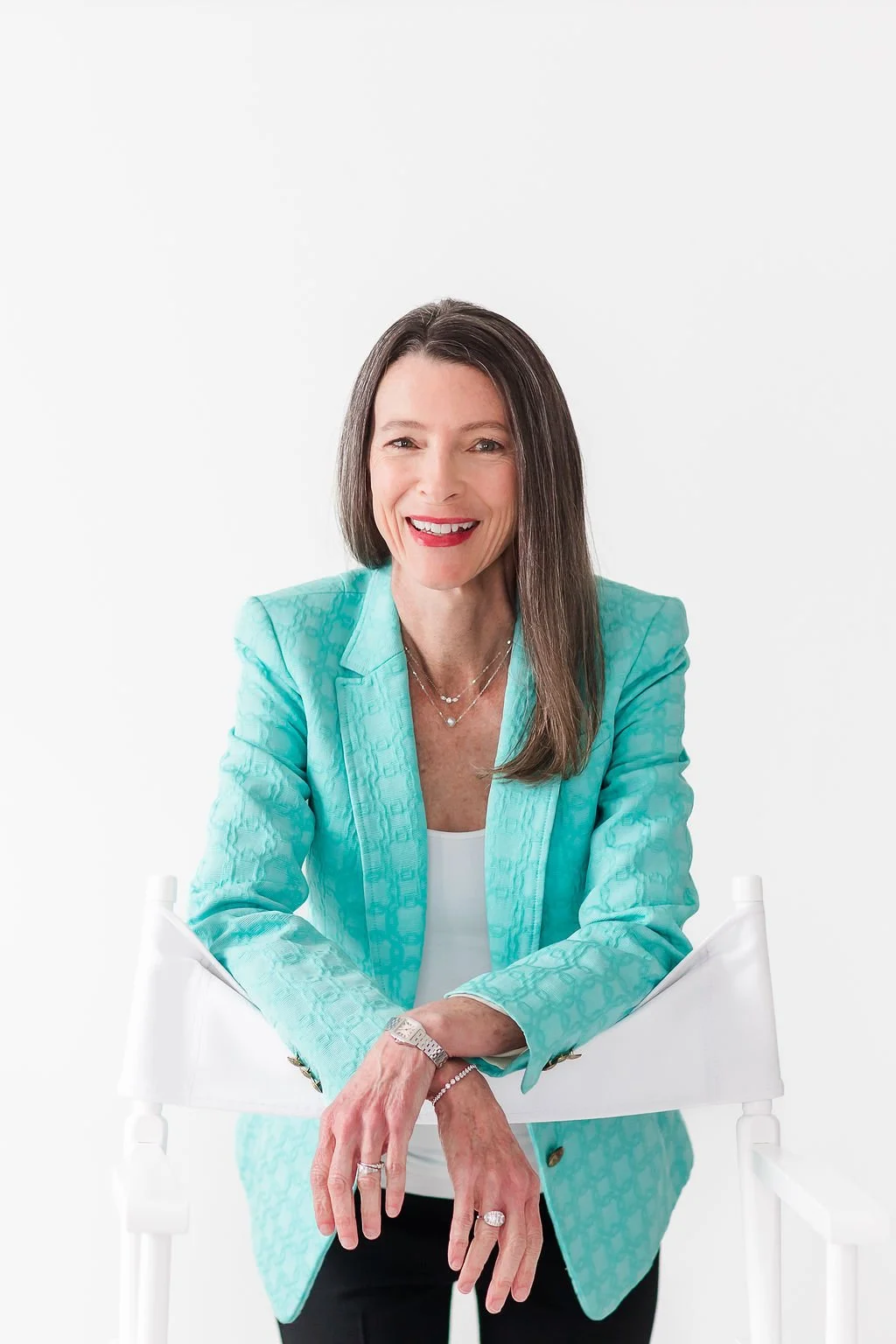 A woman with long brown hair and red lipstick sitting and smiling at the camera. She is wearing a light blue blazer over a white top, with layered necklaces and rings. The background is plain white.
