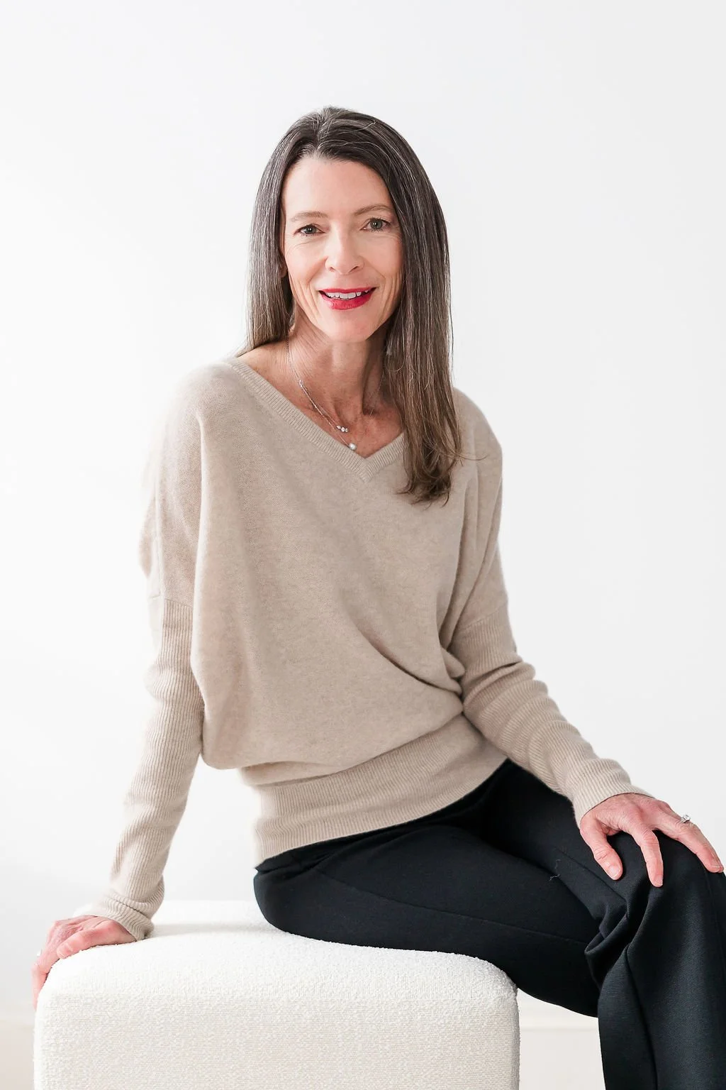 A woman with long dark hair sitting on a white ottoman against a white background, wearing a beige sweater and black pants, smiling at the camera.