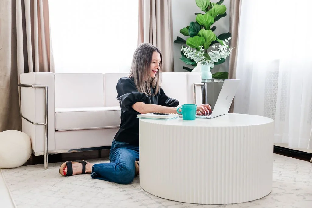 A woman kneeling on a carpeted floor in front of a white round coffee table, working on a laptop with a teal mug nearby. She is in a living room with a white sofa, a large green plant, and curtains.