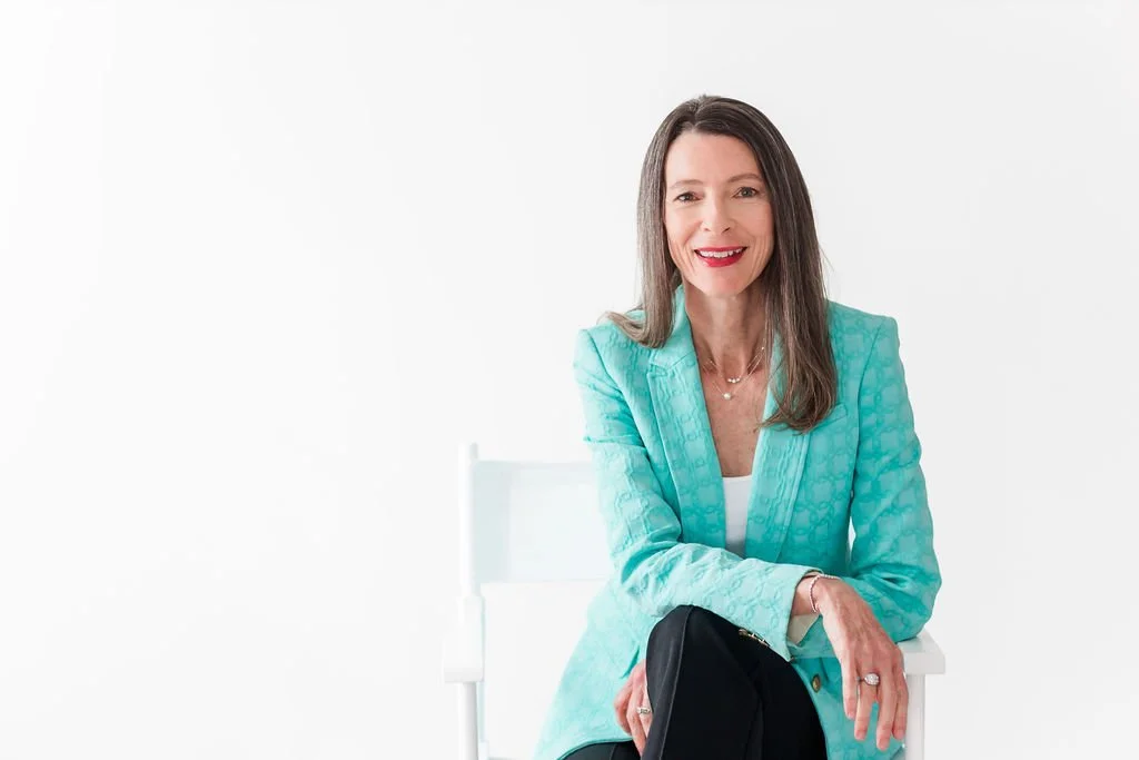 A woman with long brown hair wearing a turquoise blazer, white top, and a necklace, sitting on a white chair against a plain white background, smiling.