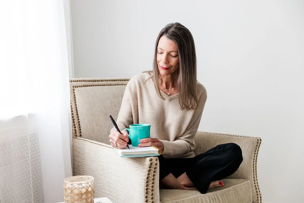 Woman sitting on a beige armchair near a window, writing in a notebook with a pen, holding a blue mug, cluttered with a candle on small table.