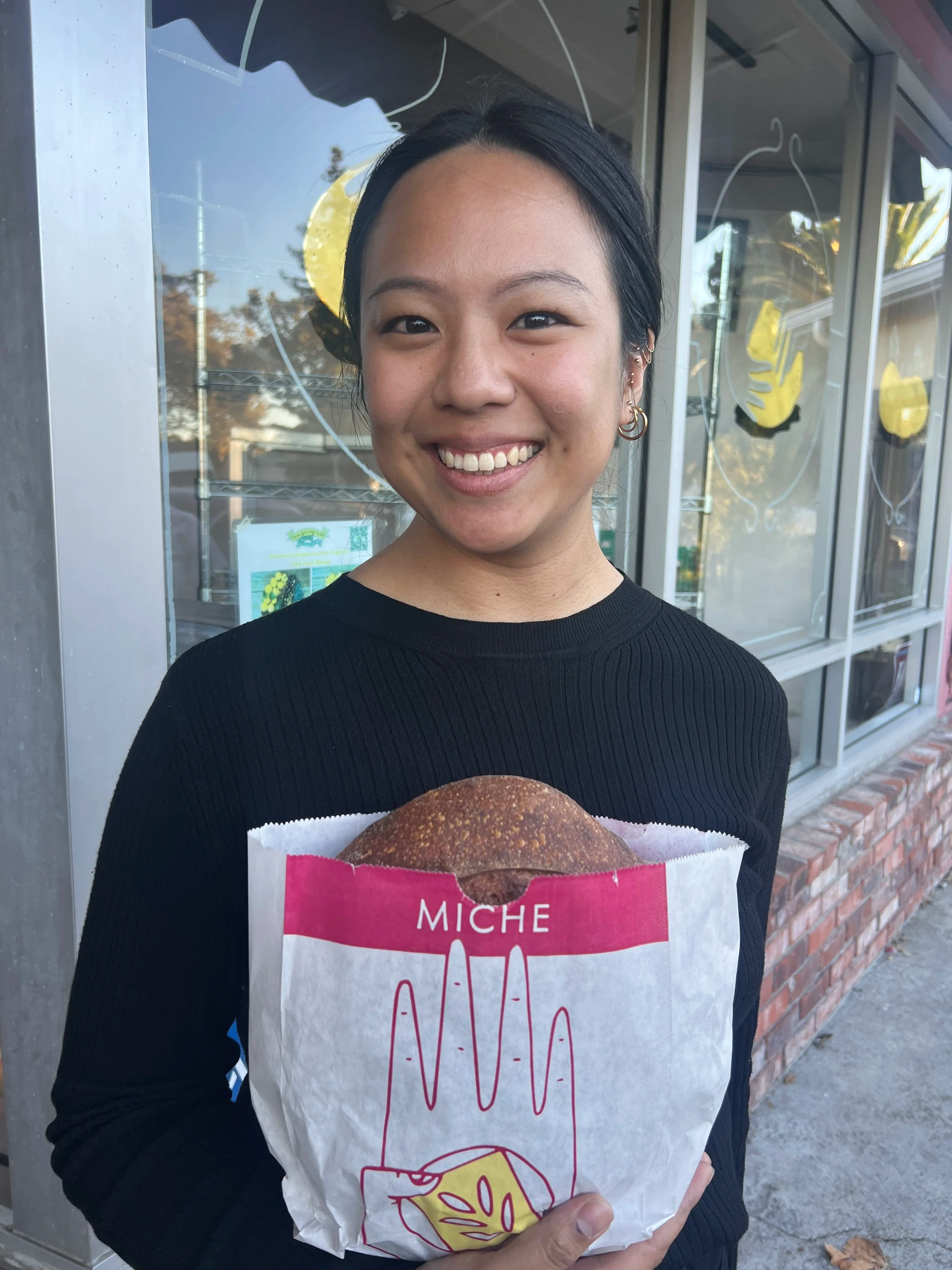 A young woman with black hair smiling and holding a wrapped sandwich or burger outside a storefront with large windows and yellow decorations.