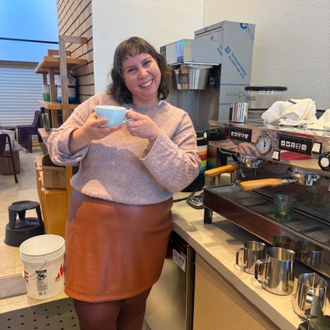 A woman smiling and holding a cup in a coffee shop or cafe, with coffee brewing equipment and supplies around her.