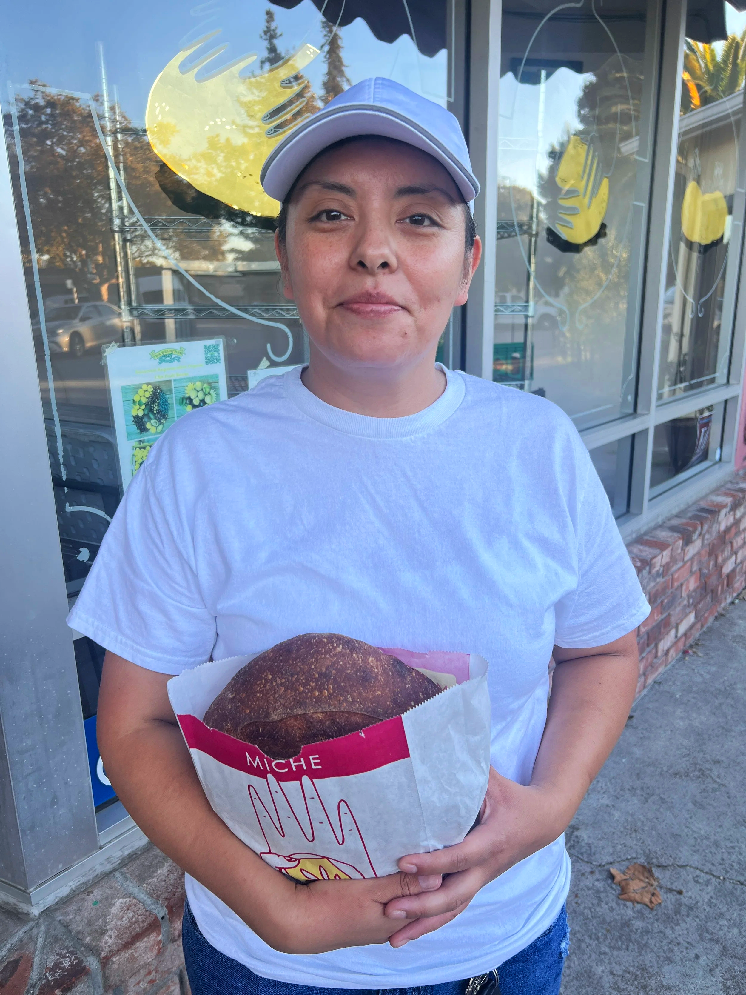 A woman wearing a white t-shirt and a light-colored baseball cap holding a paper bag with a round, dark bread loaf outside a store with large windows.