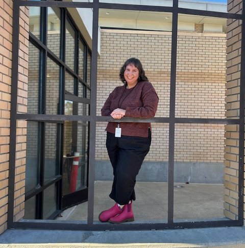 Woman in dark pants, maroon jacket, pink shoes, smiling behind a metal grid