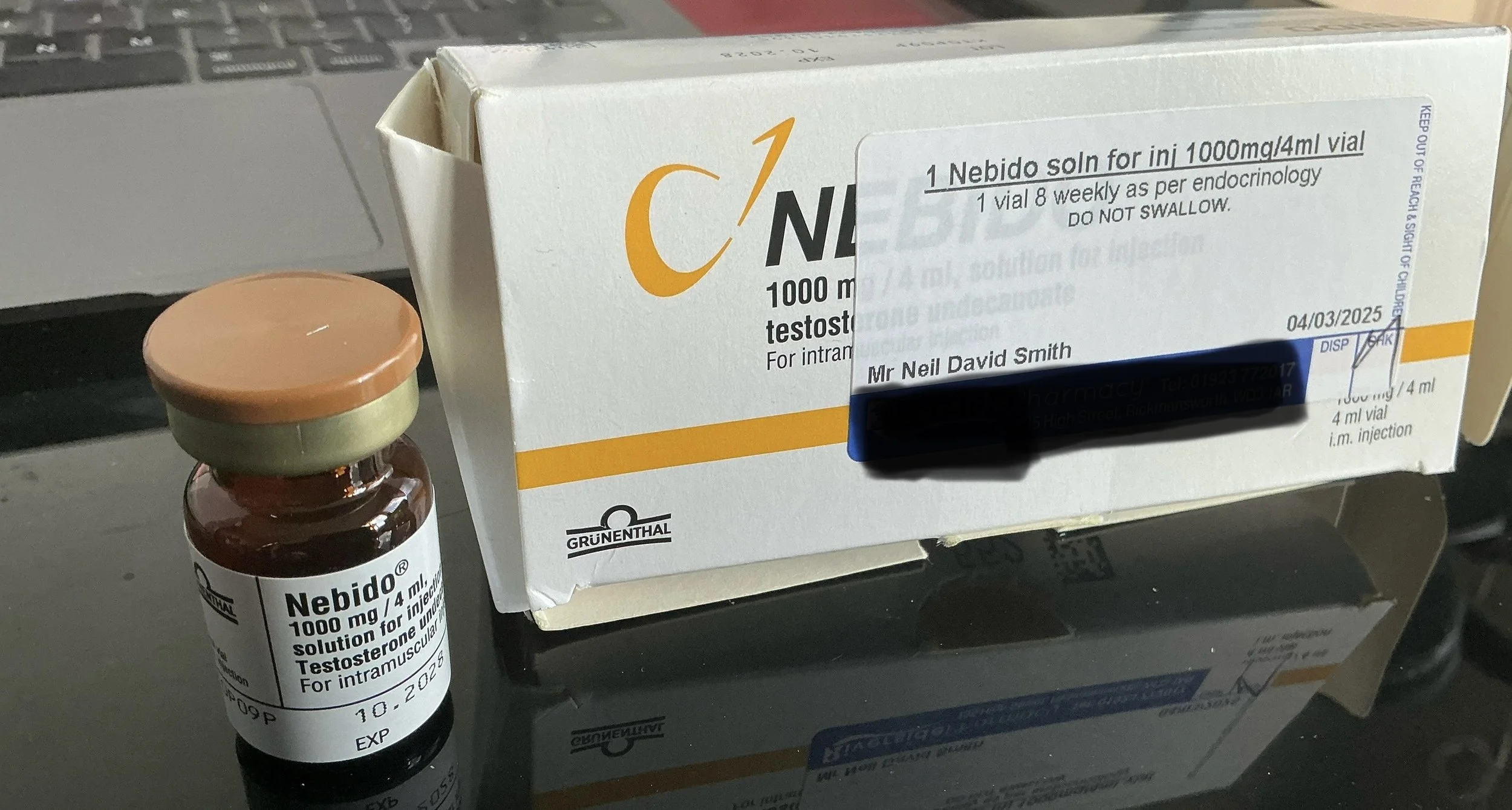 A small brown glass vial labeled 'Nebido 1000 mg/4 ml solution for injection testosterone undecanoate,' beside a white box with similar labeling. The box and vial are placed on a reflective black surface, with a laptop keyboard visible in the background.