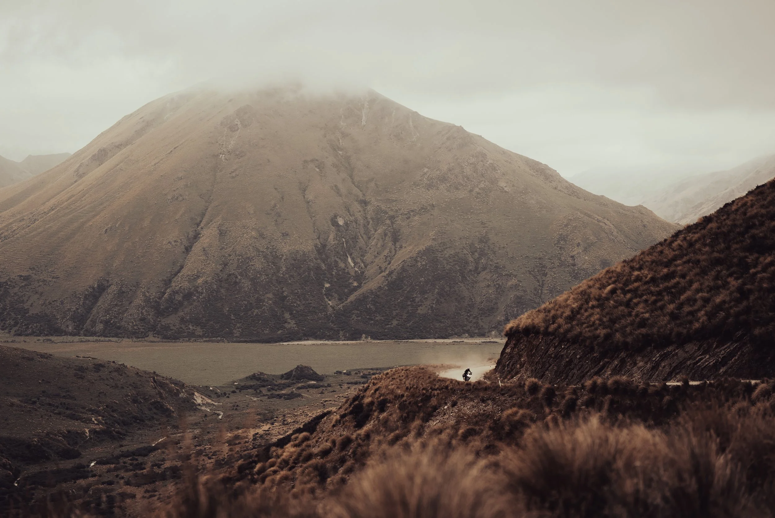 A person riding a dirt bike on a trail through a mountainous landscape with large hills and clouds around the mountain's peak.