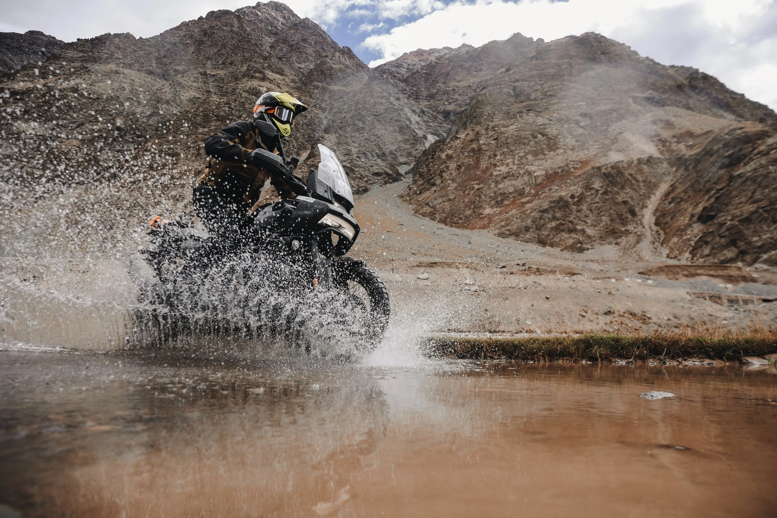Motorcycle rider wearing a helmet and riding gear crossing a shallow water body in a mountainous landscape.
