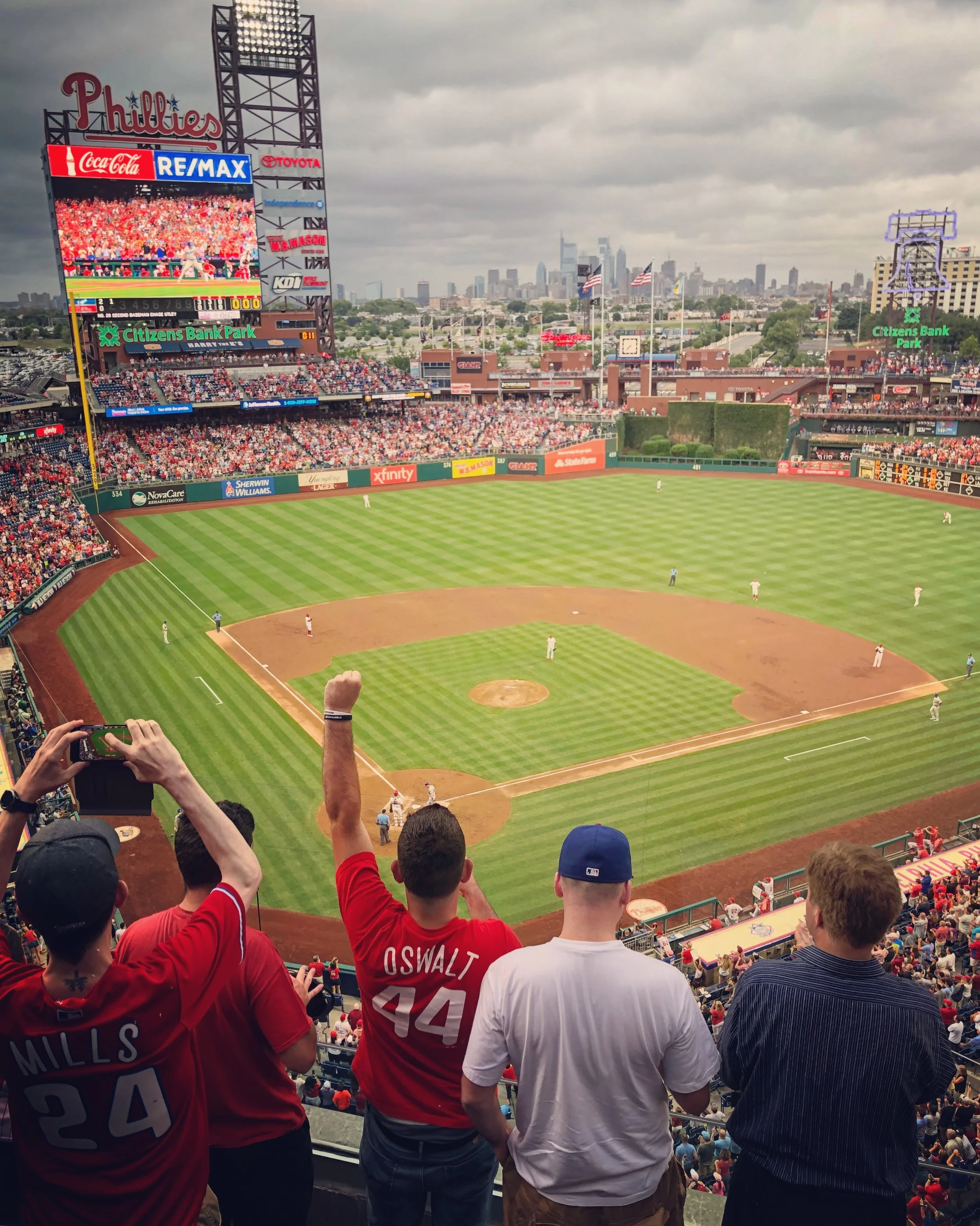 A baseball game at Citizens Bank Park stadium in Philadelphia, with fans cheering and raising their hands, and the city skyline in the background under cloudy skies.