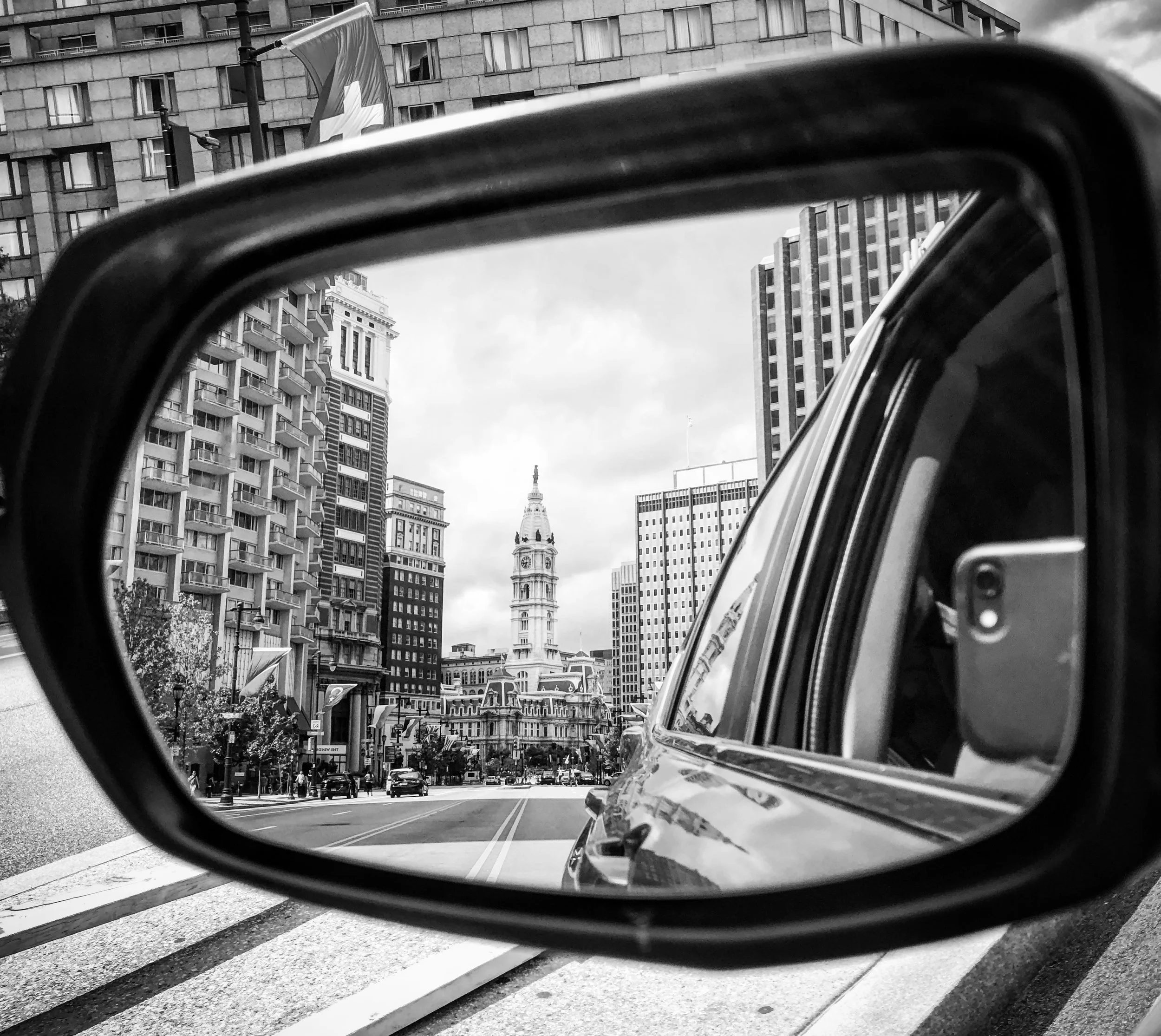 Cityscape view of a historic clock tower through a car's side mirror, surrounded by modern high-rise buildings, black and white photograph.