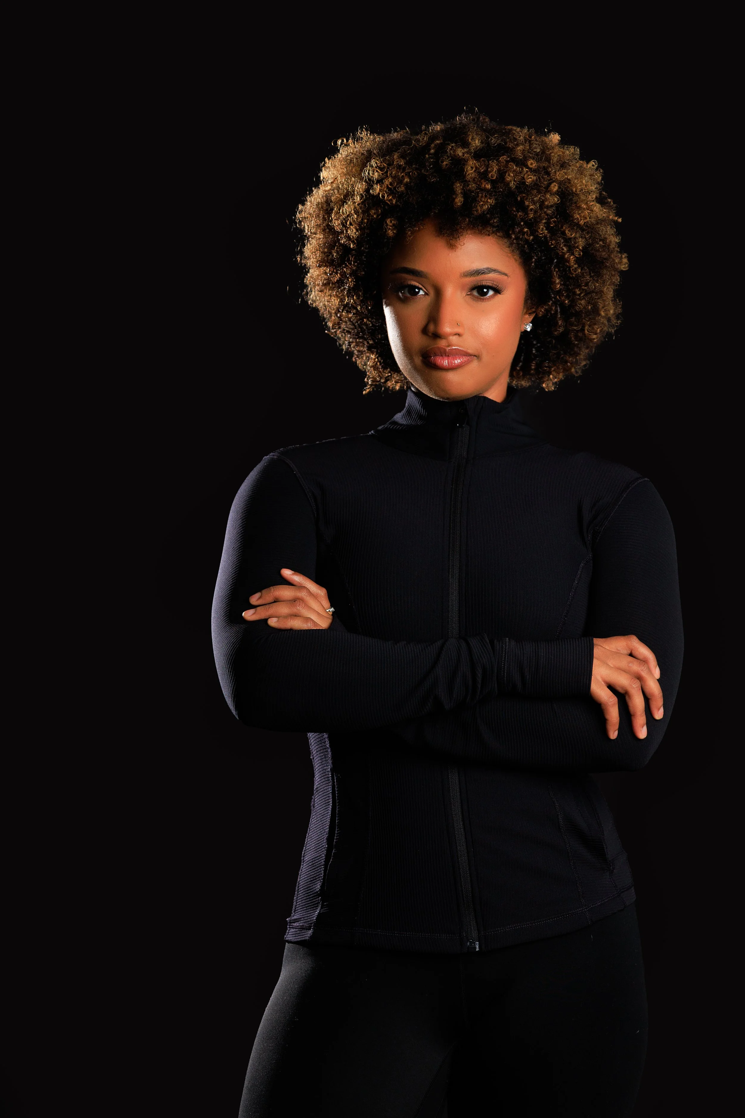 Portrait of a young woman with curly hair, crossing her arms, wearing a black athletic jacket against a black background.