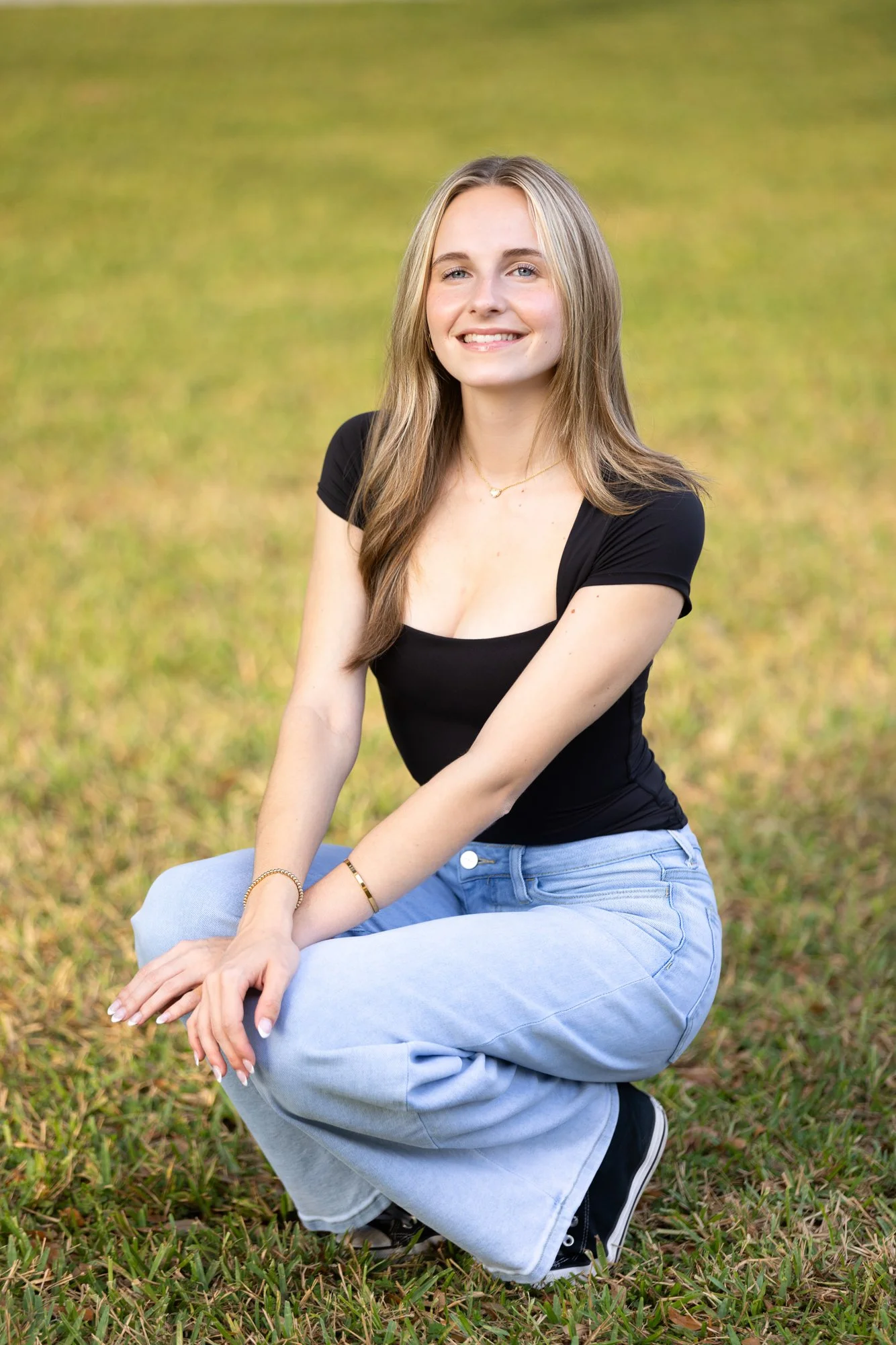 A young woman with long, light brown hair, wearing a black T-shirt and light blue baggy jeans, squatting outdoors on grass, smiling at the camera.