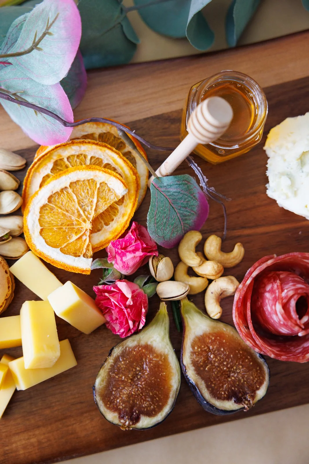 Charcuterie board with dried orange slices, cheese, figs, cashews, rose flowers, honey jar with dipper, and cured meat rolls on a wooden surface.