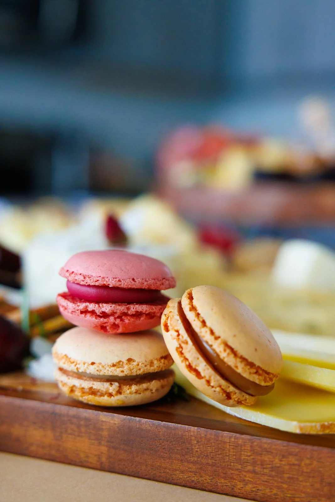 Close-up of pink and beige macarons on a wooden tray with blurred desserts in the background.