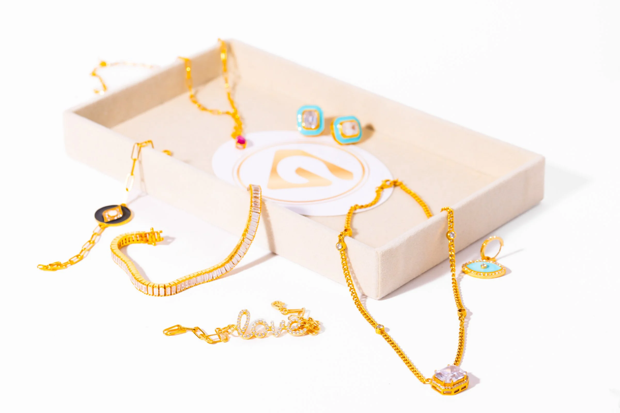 Collection of gold jewelry including necklaces, bracelets, and earrings with decorative stones, placed on a white surface and a beige jewelry box.