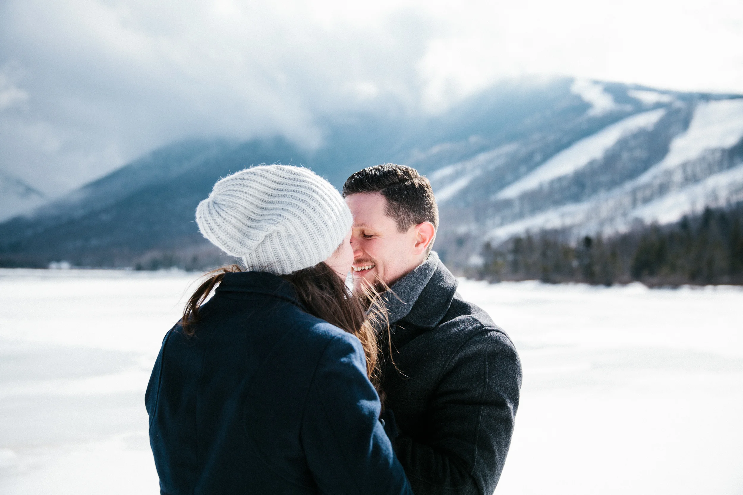 A couple in winter clothing sharing a close moment outdoors in a snowy mountain landscape.