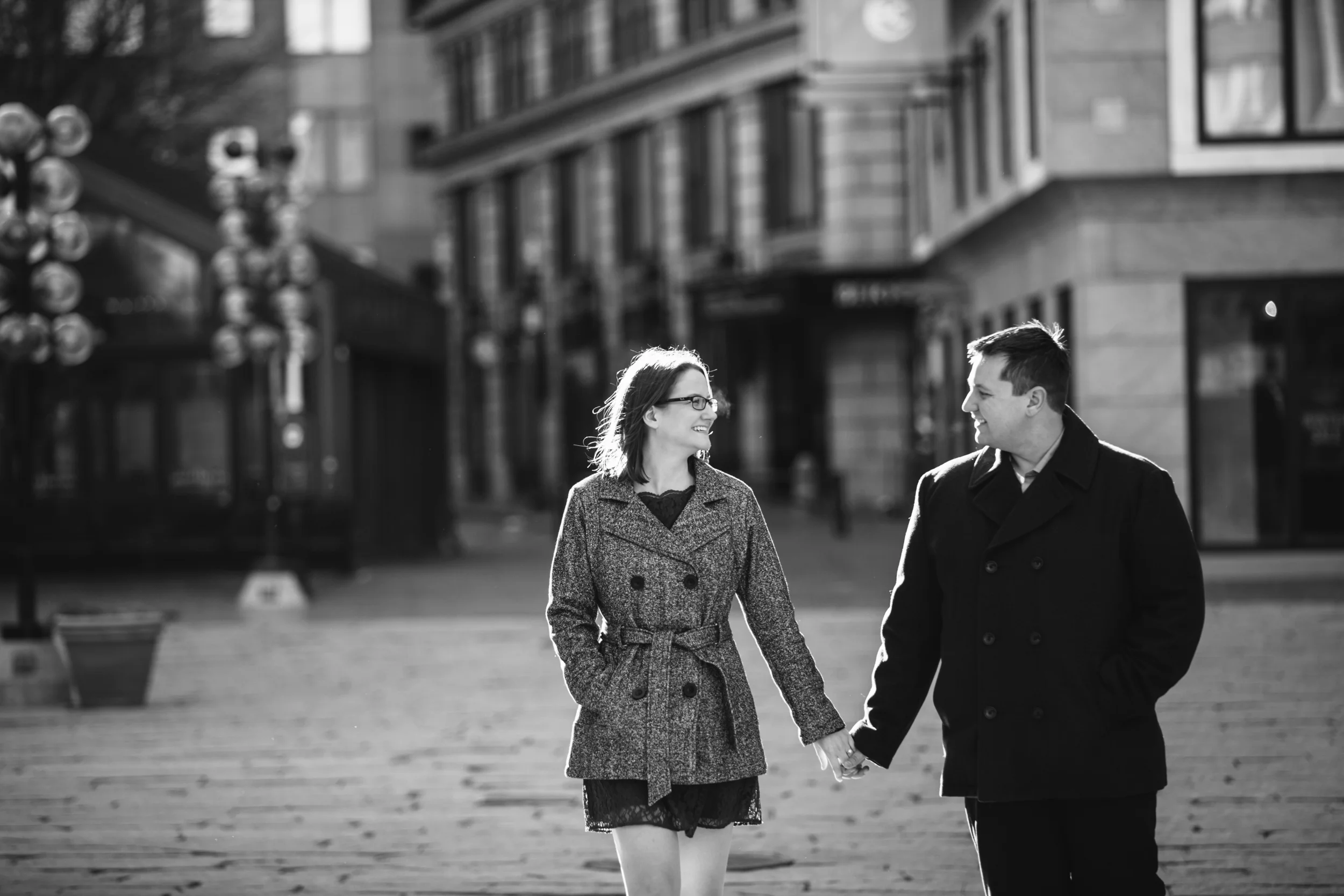 A black-and-white photo of a man and woman holding hands and smiling at each other walking on a city street.