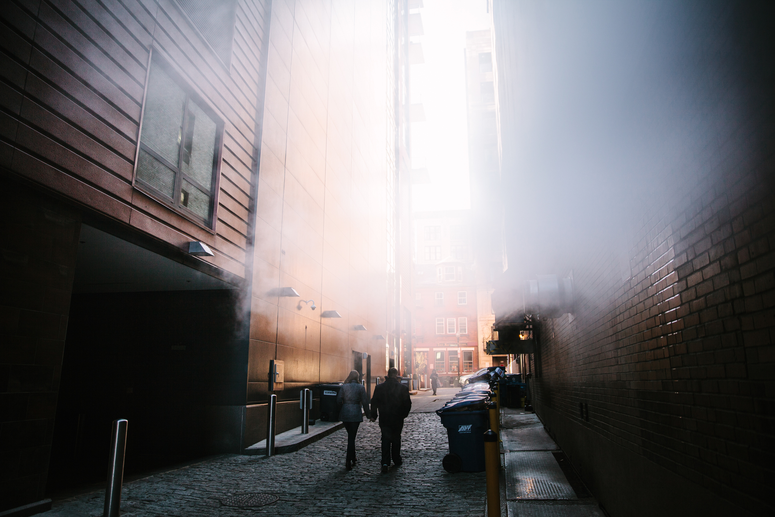 Two people walking hand in hand through a narrow cobblestone alleyway between brick and modern buildings, with smoke billowing on the right side and sunlight illuminating the alley.